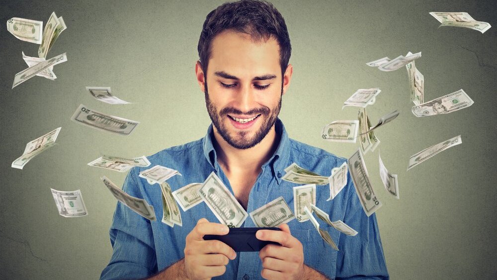 Technology online banking money transfer, e-commerce concept. Happy young man using smartphone with dollar bills flying away from screen isolated on gray wall office background.