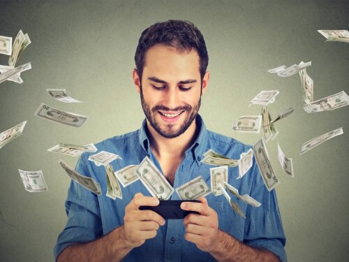 Technology online banking money transfer, e-commerce concept. Happy young man using smartphone with dollar bills flying away from screen isolated on gray wall office background.